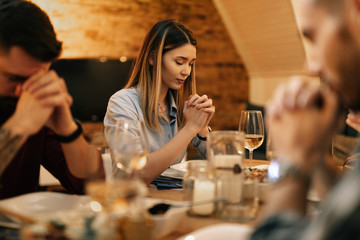 Young woman and her friends praying before a meal at dining table.