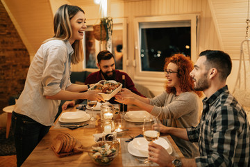 Young cheerful woman serving food to her friends while having dinner in dining room.