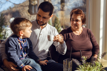Caucasian family small child boy son three years old sitting in a lap of his father or uncle young man holding a cup of coffee while senior woman grandmother is looking grandson by table at cafe