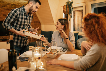 Young happy man serving dinner to his friends in dining room.