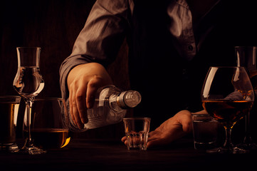 The bartender pours the vodka or tequila in small shot glass on the old bar counter. Vintage wooden background in pub or bar, night mood. Place for text, toning, selective focus