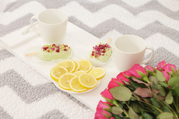 a white wooden tray with two cups, two cakes in the form of popsicle, a plate with cut lemon and a bouquet of pink roses on the bed..