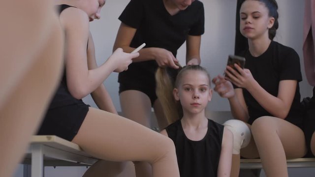 Medium shot of teen and preteen girls hanging out in locker room before cheerleading or gymnastics practice. One of them is making ponytail for her friend while others looking at mobile phones