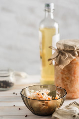 Fermented cabbage in a transparent plate, in the background a glass jar with cabbage, a bottle of oil, with a wooden spoon and a linen napkin on a light background.