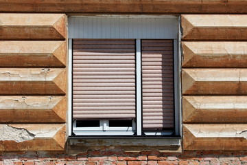 Fototapeta na wymiar Renovated double window with white wooden frame and plastic blinds mounted on cracked dilapidated broken striped with horizontal lines old style decorative facade above suburban family house red brick
