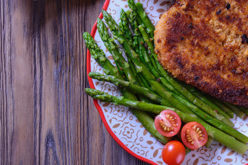 Appetizing schnitzel with crispy crust on a plate next to asparagus, cherry tomatoes  on a wooden...