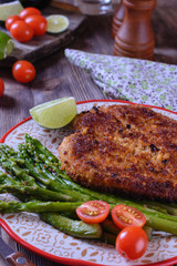 Appetizing schnitzel with crispy crust on a plate next to asparagus, lime and cherry tomatoes on a wooden table are ready for eating, vertical orientation