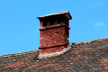 Single elongated narrow old dark red bricks dilapidated cracked chimney with multiple openings covered with small roof tiles on top of suburban family house roof