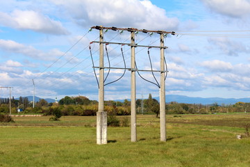 Three connected large concrete power line utility poles with multiple electrical wires connected with glass insulators surrounded with uncut grass and trees on cloudy blue sky background