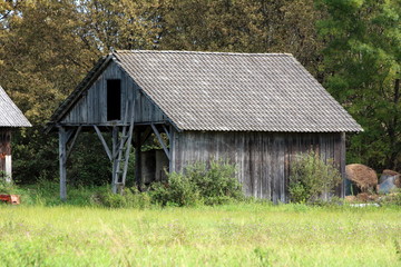 Fototapeta premium Vintage retro rustic old dilapidated wooden barn made from wooden boards with open area filled with hay bales for storage over winter surrounded with uncut grass and dense trees in background