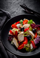 Fresh salad with chicken breast, cheese, black olives,red pepper, lettuce, fresh sald leaves and tomato on a black plate on Dark grey black slate background