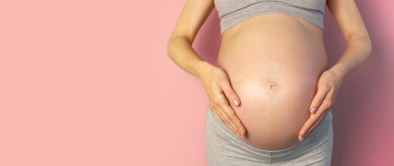 Beautiful young pregnant woman holds hands on her belly on pink background. The concept of expectation of the child, pregnancy and motherhood. Tender photo of a pregnant. Close-up, place for text