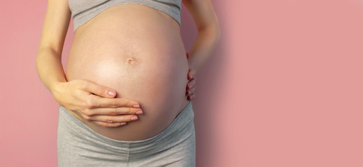 Beautiful young pregnant woman holds hands on her belly on pink background. The concept of expectation of the child, pregnancy and motherhood. Tender photo of a pregnant. Close-up, place for text - obrazy, fototapety, plakaty