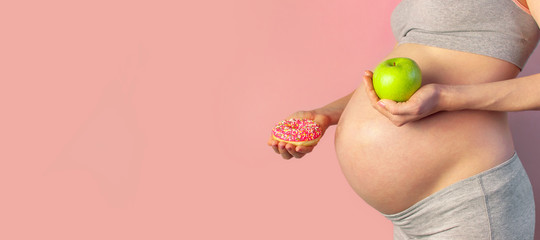 Beautiful young pregnant woman holding green apple and sweet donut in her hand on pink background. Expectation of the child, pregnancy and motherhood. Healthy and unhealthy eating concept, diet