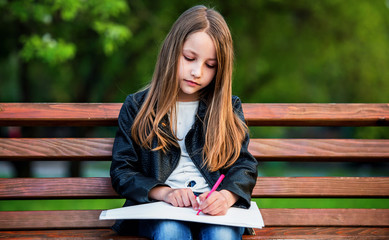 Fototapeta na wymiar Little girl sitting on the park bench and drawing with a felt pen. Lifestyle, childhood, education concept
