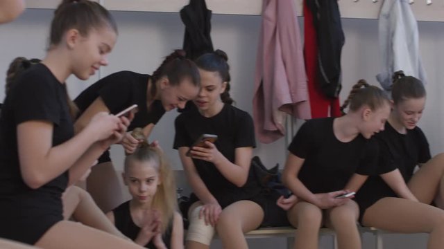 Tracking shot of teen cheerleading team getting ready for practice in locker room. They are chatting and putting on gymnastic shoes. One of them is making ponytail