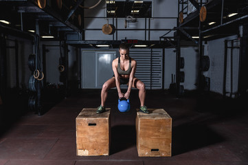 Young sweaty muscular fit girl with big muscles and strong legs doing deep squat and dip on two wooden jump boxes with heavy kettlebell for hardcore crossfit strength workout training in the gym 