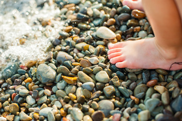 Child's feet on a beach in small color pebble in a summer sunny day. Holiday on a seashore.