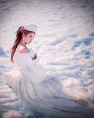 Fototapeta na wymiar Amazing cute slim young woman in chic fairy tale image in white puffy dress and with crown on her head sits on snow in sunny winter day