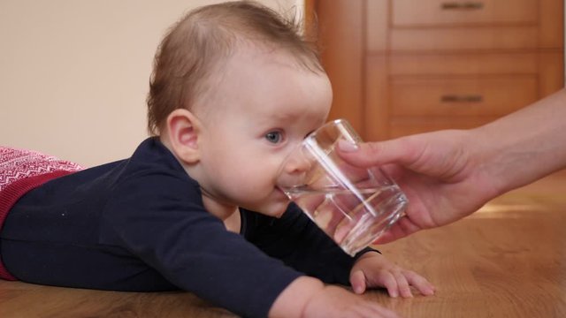 Mother gives baby child a glass of water and child drinks it lying on floor at home