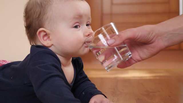 Mother gives baby child a glass of water and child drinks it lying on floor at home