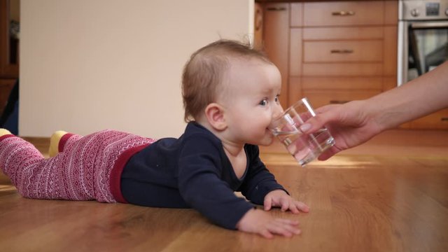 Mother gives baby child a glass of water and child drinks it lying on floor at home