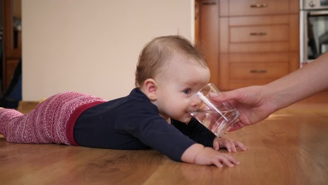 Mother gives baby child a glass of water and child drinks it lying on floor at home