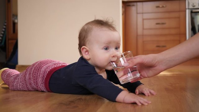 Mother gives baby child a glass of water and child drinks it lying on floor at home