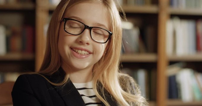 Close up of blonde Caucasian pretty teen small girl in glasses finishing to read book and closing it. Beautiful little blond schoolgirl smiling to camera in library. Portrait of cute smiled kid.