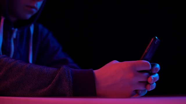 Young man in a hood with a smartphone. Hacker makes a hack through the phone. Blue and red light falls on a man on a black background.