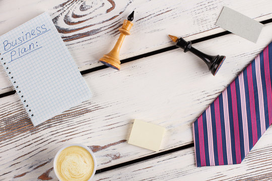 Office man flatlay. Notebook with business plan, chess figures and paper stuff. Cup of cappuccino and striped tie.