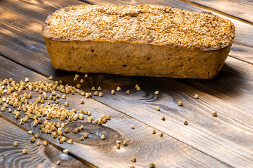 Gluten-free buckwheat bread with a golden brown crust, sprinkled with sesame seeds, lies on a wooden table. Healthy homemade recipe. Grains of green buckwheat are scattered nearby. Copy space © Jekh