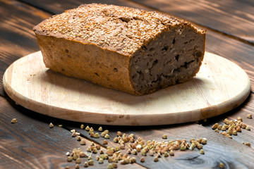 A loaf of freshly baked buckwheat bread lies on a round kitchen board on a brown wooden table. A handful of green buckwheat seeds are scattered nearby. Harmless baking for vegans and vegetarians