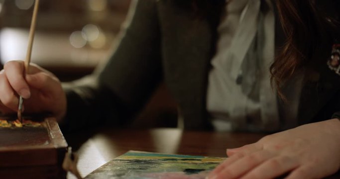 Close up of Caucasian teen hands of girl painting picture with brush and oil paints at desk in library. Schoolgirl drawing canvas with colors in dark clasroom.