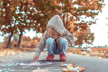 Little boy draws with colored crayons on asphalt drawings, concept creative art and creativity in teenage period. Background trees in park in autumn. Casual warm clothes with a hood.