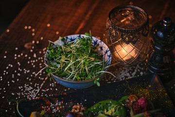 Healthy appetizer of a tiny bowl of greens and herbs, next to salt grinder, decorated with spices on a wooden table in a warm light of a candle in restaurant, close up view from the side