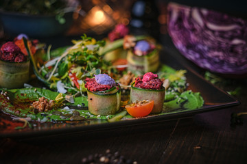 Fototapeta na wymiar Delicious appetizer of grilled and oven baked cucumber rolls served with vegan spread, thin sliced vegetables, cherry tomatoes and oil sauce, on a black porcelain dish next to a tiny bowl of greens