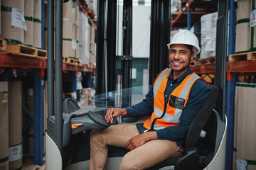 Happy young african forklift manager transporting goods from one shelf to another while looking in...