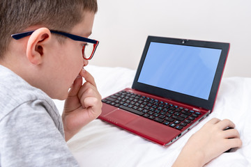 A boy wearing glasses lying in bed and using a laptop, typing the text on a keyboard close up, online education concept