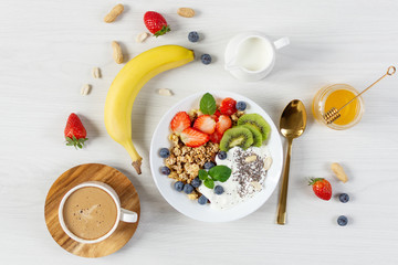 Bowl of granola with yogurt, fresh berries and fruits, cup of coffee on white wooden table background top view.