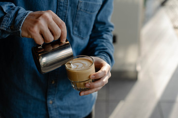 Young professional barista pouring hot milk in cup of latte. Close up of man hands drawing flower...