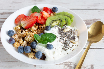 White ceramic bowl of homemade granola with yogurt, fresh berries and fruits on white wooden table background.