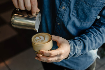 Young professional barista pouring hot milk in cup of latte. Close up of man hands drawing flower on cappuccino in modern coffee shop