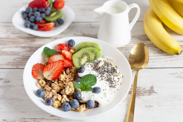 White ceramic bowl of homemade granola with yogurt, fresh berries and fruits on white wooden table background.