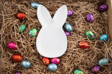 Selection of colorful foil wrapped Easter eggs scattered on shredded packing paper with white porcelain bunny shape dish in the middle. Sustainability concept at Easter. Rustic, recycled paper look.