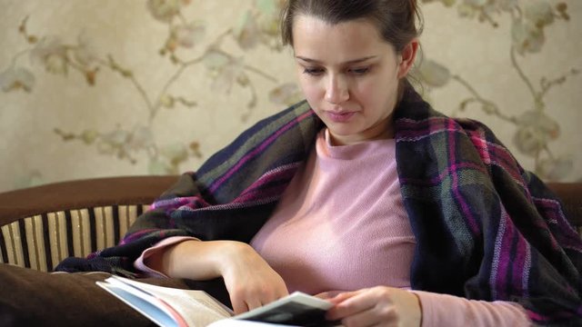 The average plan of nice and thoughtful girl sits at sofa and reads a book. She has brown blanket on her shoulders. Woman got cold. She suts at window