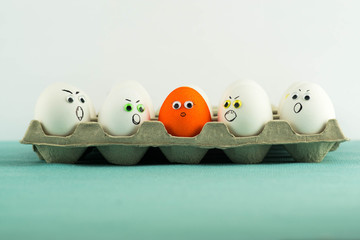 Paper tray full of angry white eggs and only one different orange egg with woman face and long...
