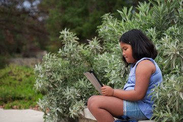 A young girl wearing jean shorts that is sitting on a bench using her wireless tablet to surf the internet while waiting.