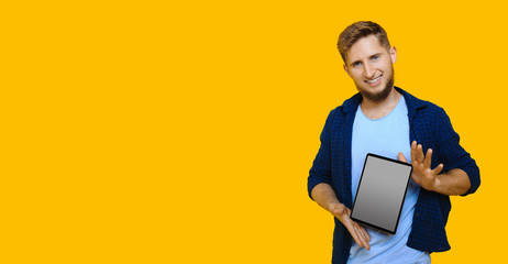 Bearded caucasian man with red hair is advertising something using a tablet with blank space on a yellow wall