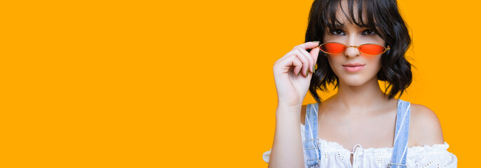 Stunning caucasian lady with black curly hair is touching her sunglasses and posing on a yellow background with blank space
